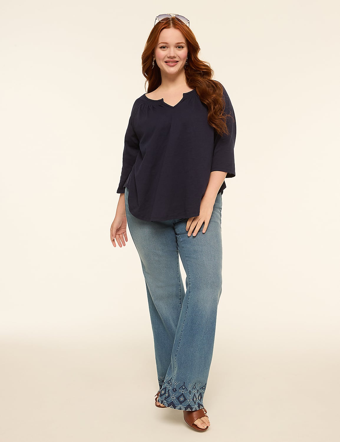 A smiling woman with long red hair stands in the foreground wearing a navy blue top and light wash jeans with embroidered hems, against a plain, light-colored background. Product Image 3