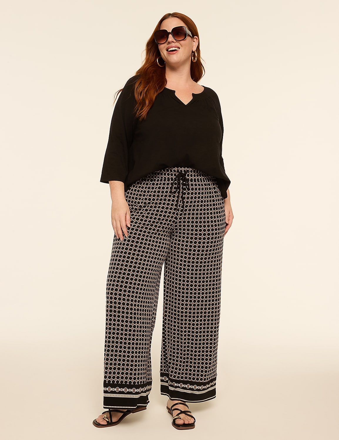 A woman with auburn hair and sunglasses stands in the foreground wearing a black top and patterned wide-leg pants against a plain, light-colored background. Product Image 3