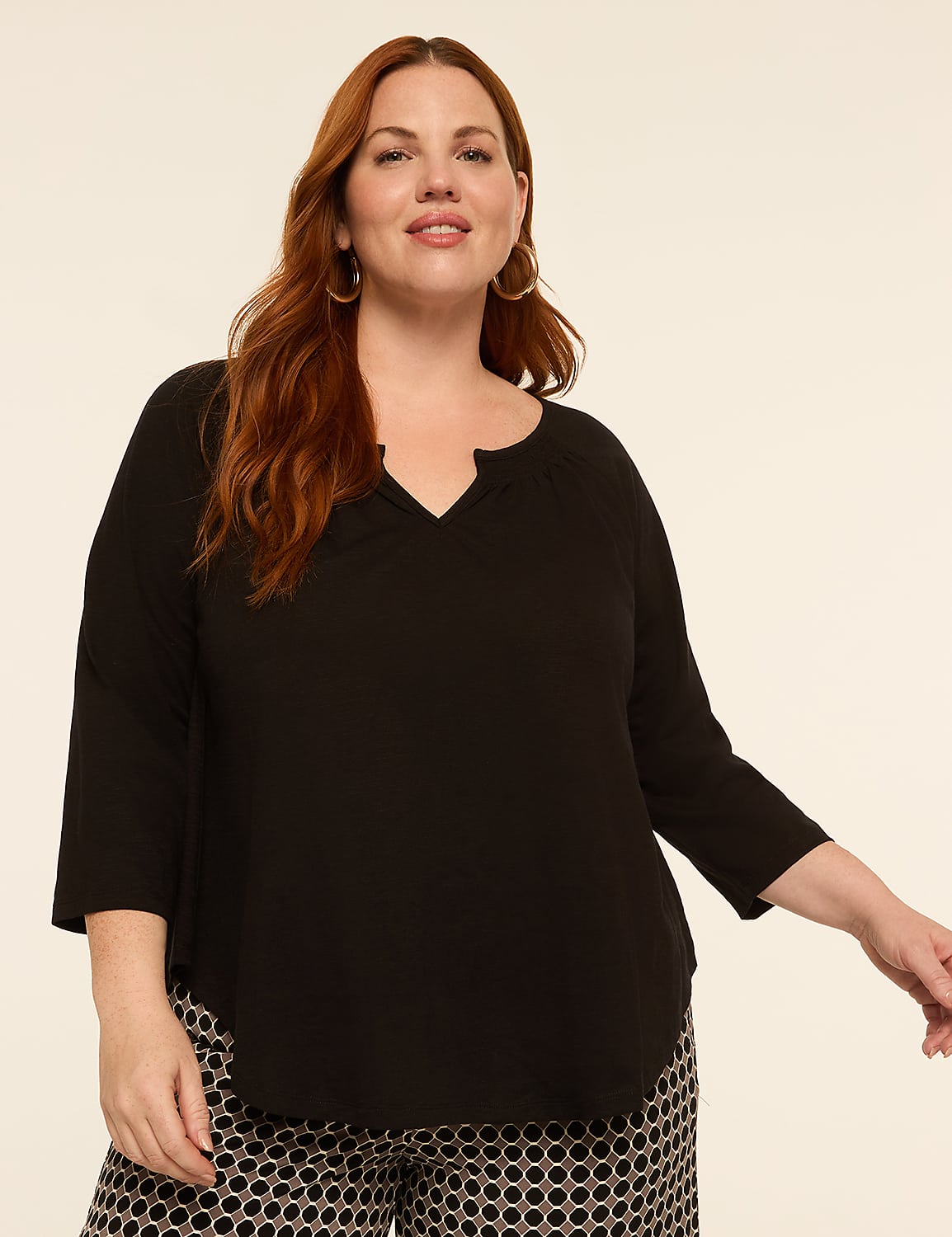 A woman with auburn hair and hoop earrings smiles while wearing a black top and patterned pants against a plain background. Product Image 1