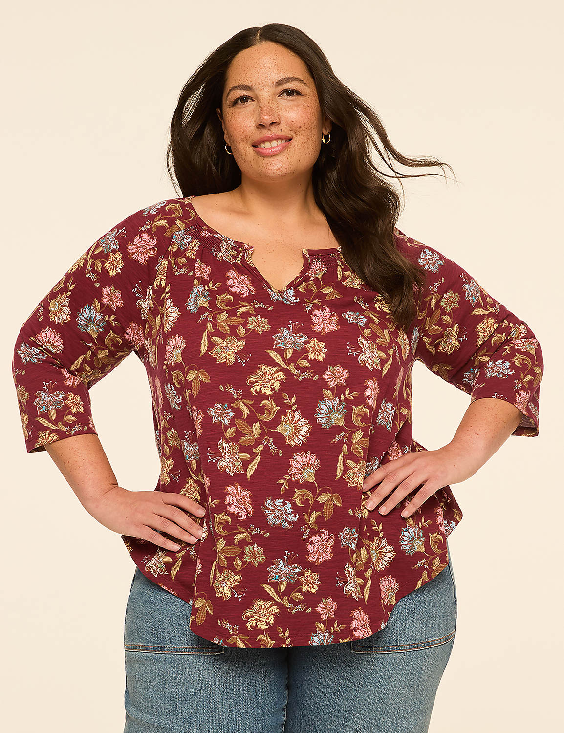 A woman with dark hair and freckles smiles while wearing a floral-patterned maroon top and blue jeans against a plain beige background. Product Image 1