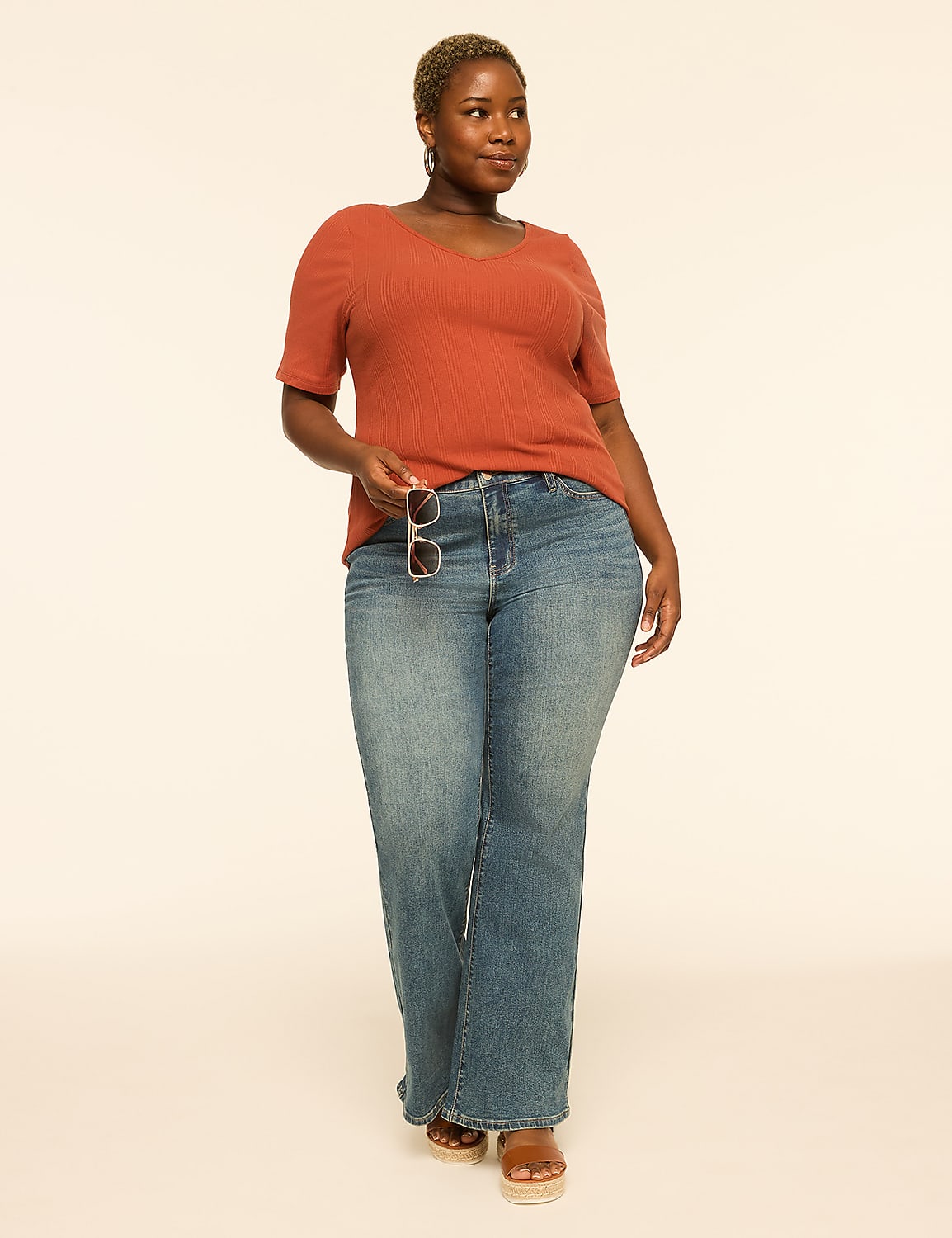 A woman in a rust-colored top and blue jeans stands against a plain, light-colored background, holding sunglasses. Product Image 3