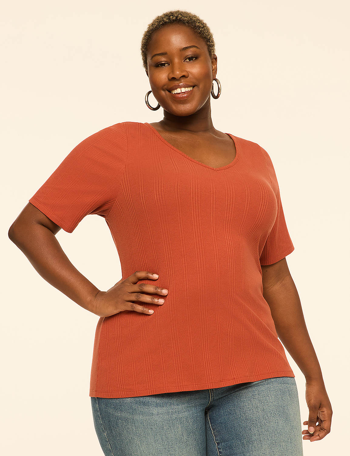 A smiling woman in a rust-colored ribbed v-neck shirt and blue jeans stands against a plain cream background. Product Image 1
