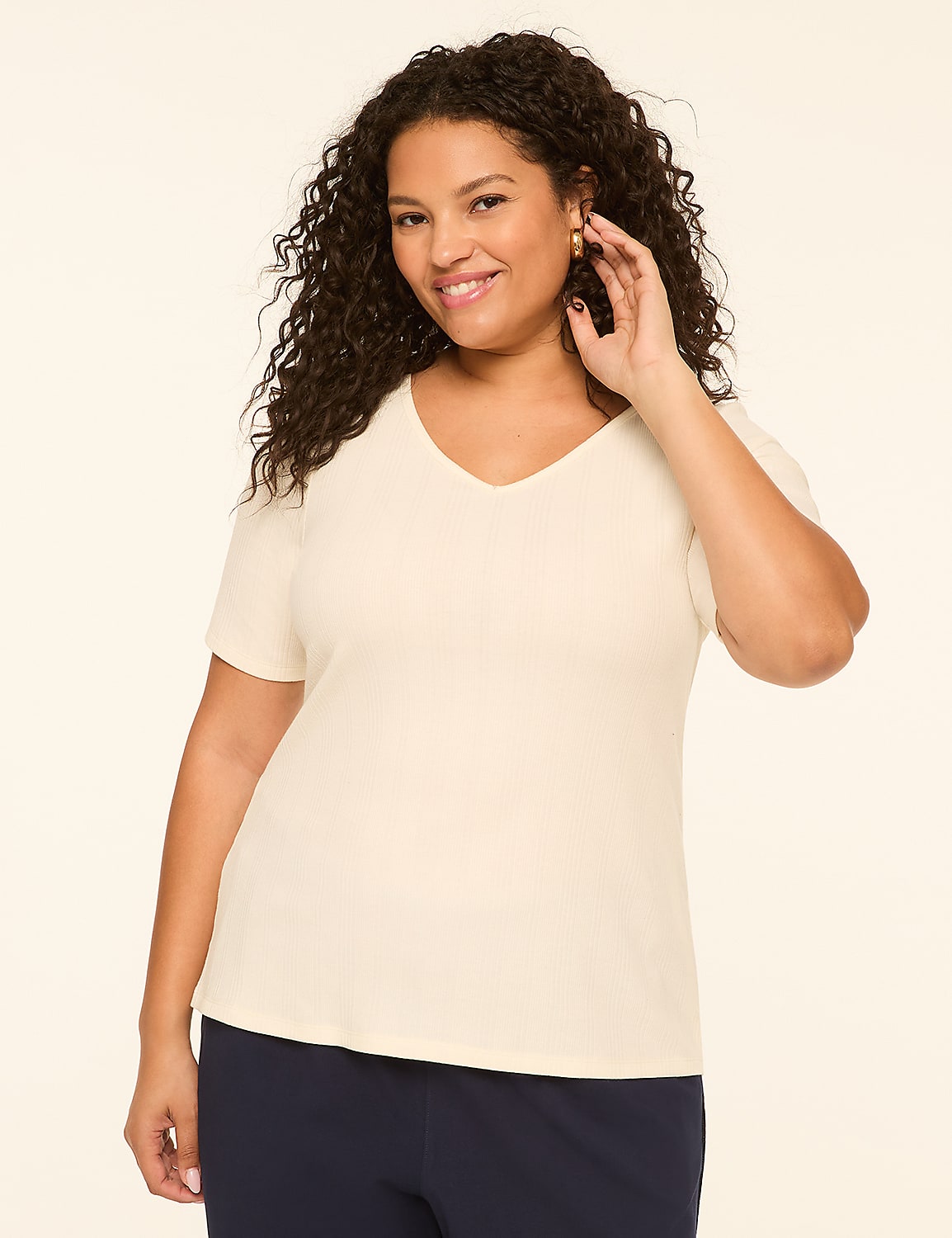A smiling woman with curly dark hair in a cream v-neck shirt and navy pants stands against a plain cream background. Product Image 1