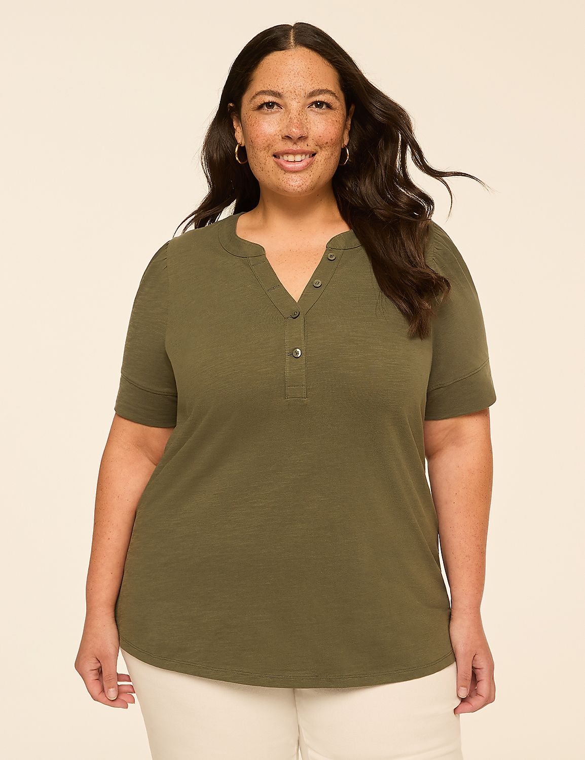 A smiling woman with dark hair and fair skin, wearing a green henley shirt and white pants, stands against a plain, light-colored background. Product Image 1