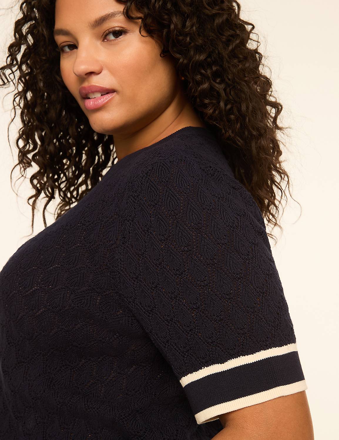 A woman with curly dark hair and a navy blue knit top with white stripes on the sleeves is shown against a plain, light-colored background. Product Image 4
