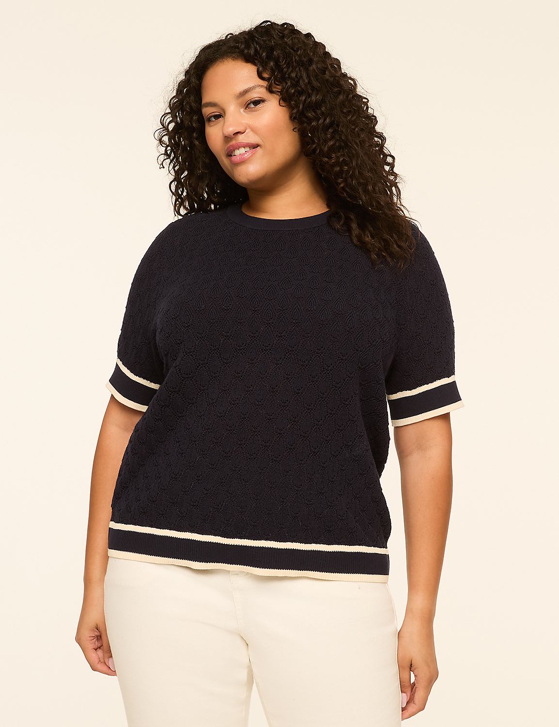 A plus-size woman with curly dark hair poses in a navy blue short-sleeved sweater with cream trim against a plain cream background. Product Image 1