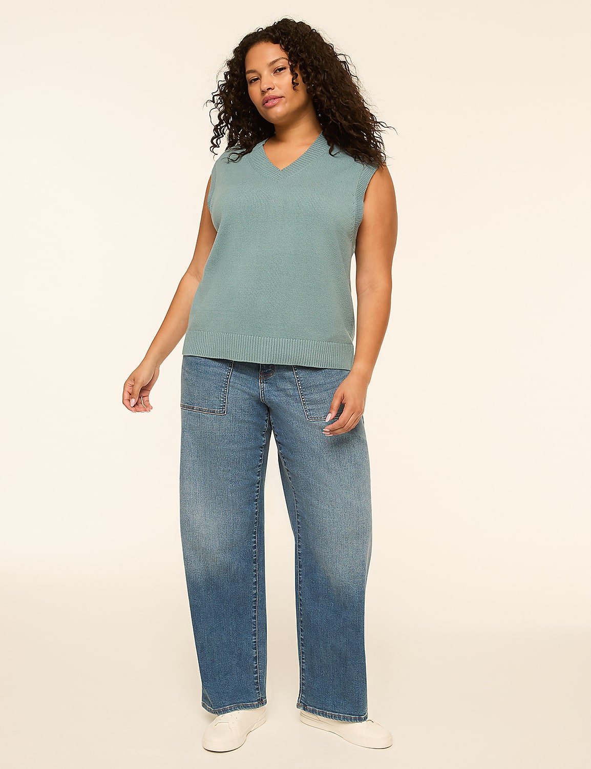 A woman in a teal v-neck sweater vest and baggy blue jeans stands against a plain cream background. Product Image 1