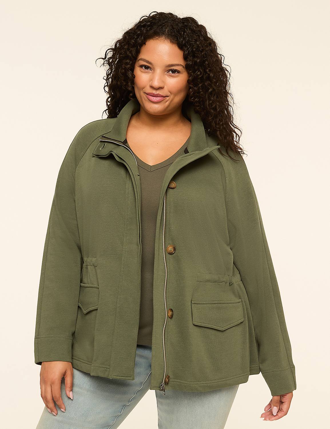 A smiling woman with curly dark hair stands in the foreground wearing a green jacket and light blue jeans, against a plain, light beige background. Product Image 1