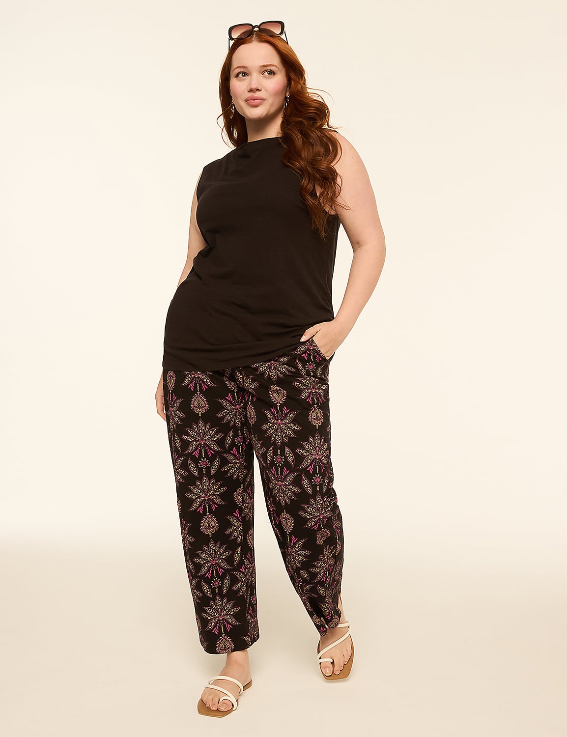 A woman with red hair stands in the foreground wearing a black sleeveless top and patterned pants, against a plain, light-colored background. Product Image 3