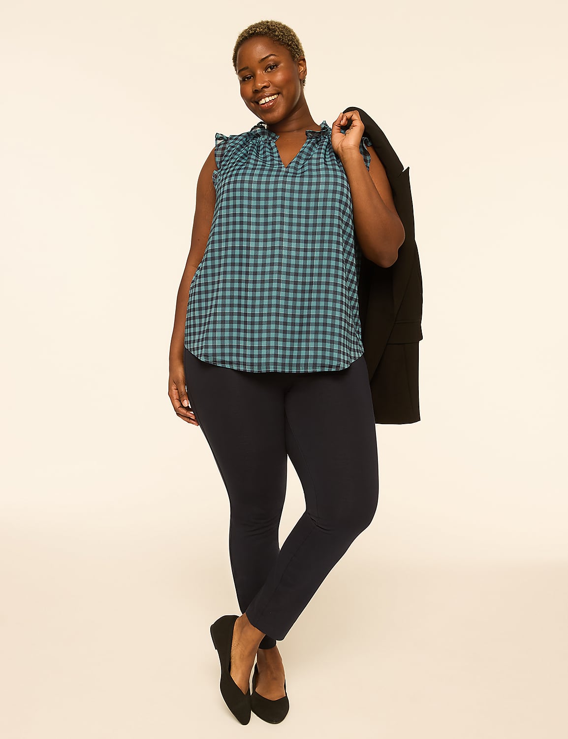 A smiling woman in a blue gingham top and dark pants stands against a plain, light-colored background, with a black jacket draped over her shoulder. Product Image 3