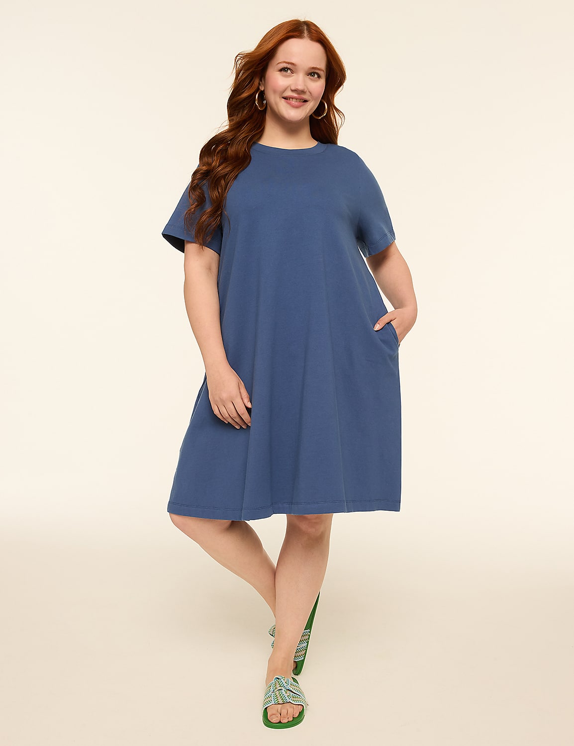 A woman with long, wavy red hair stands with one leg crossed in front of the other, wearing a blue t-shirt dress and green sandals against a plain, light-colored background. Product Image 1