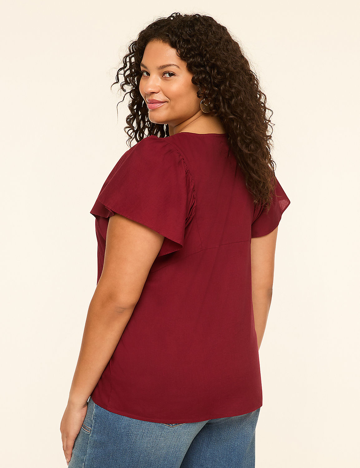 A woman with curly dark hair in a maroon top and blue jeans stands against a plain cream background. Product Image 2