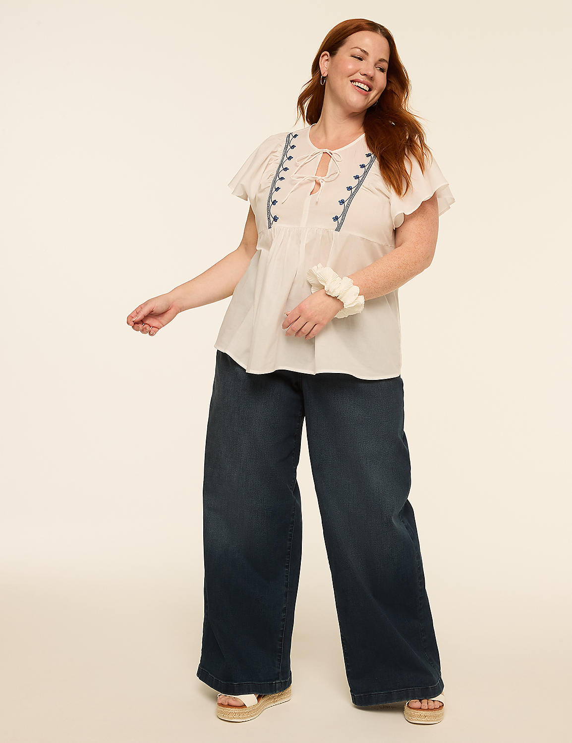 A woman with auburn hair smiles and poses in a white embroidered top and wide-leg jeans against a plain, light background. Product Image 3