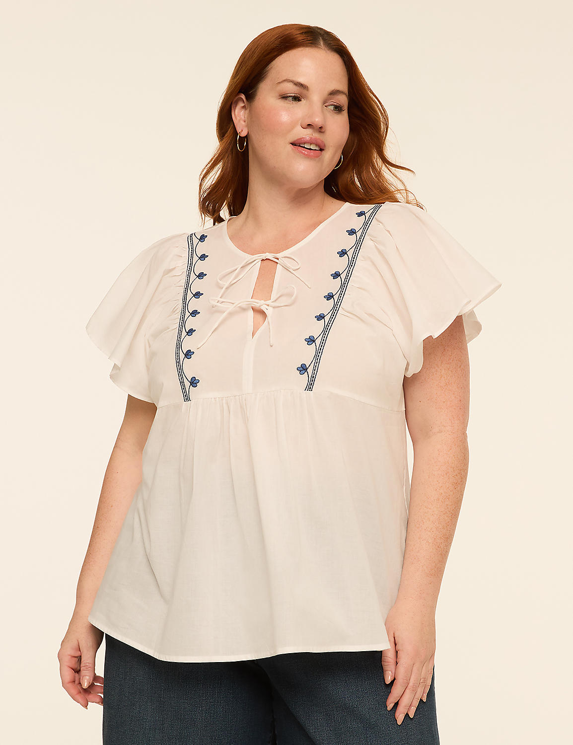A woman with auburn hair and fair skin, wearing a white embroidered top with flutter sleeves, stands against a plain, light-colored background. Product Image 1