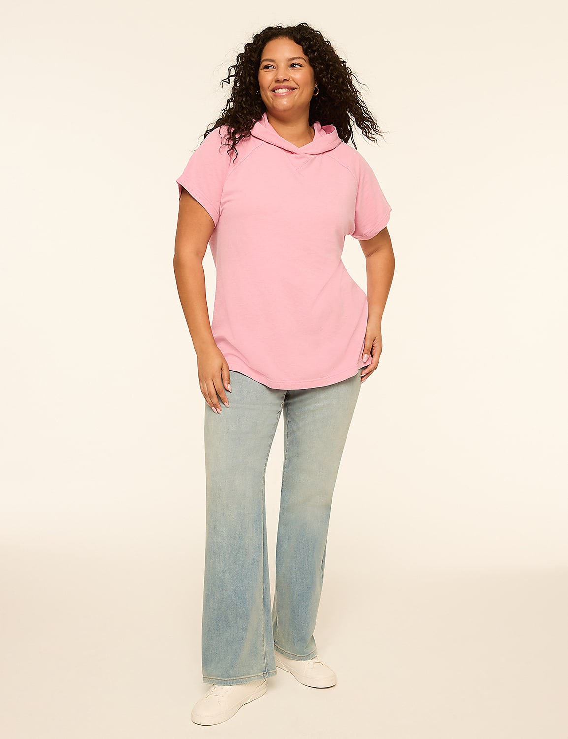 A smiling woman with curly dark hair stands in the foreground wearing a pink hooded shirt and light-wash flared jeans against a plain, light-colored background. Product Image 3