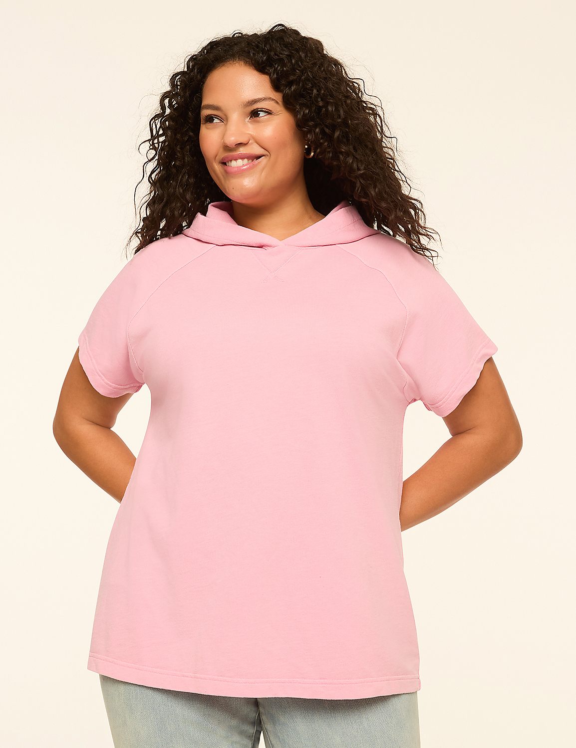A smiling woman with curly dark hair stands in the foreground wearing a pink short-sleeved hoodie against a plain, light-colored background. Product Image 1