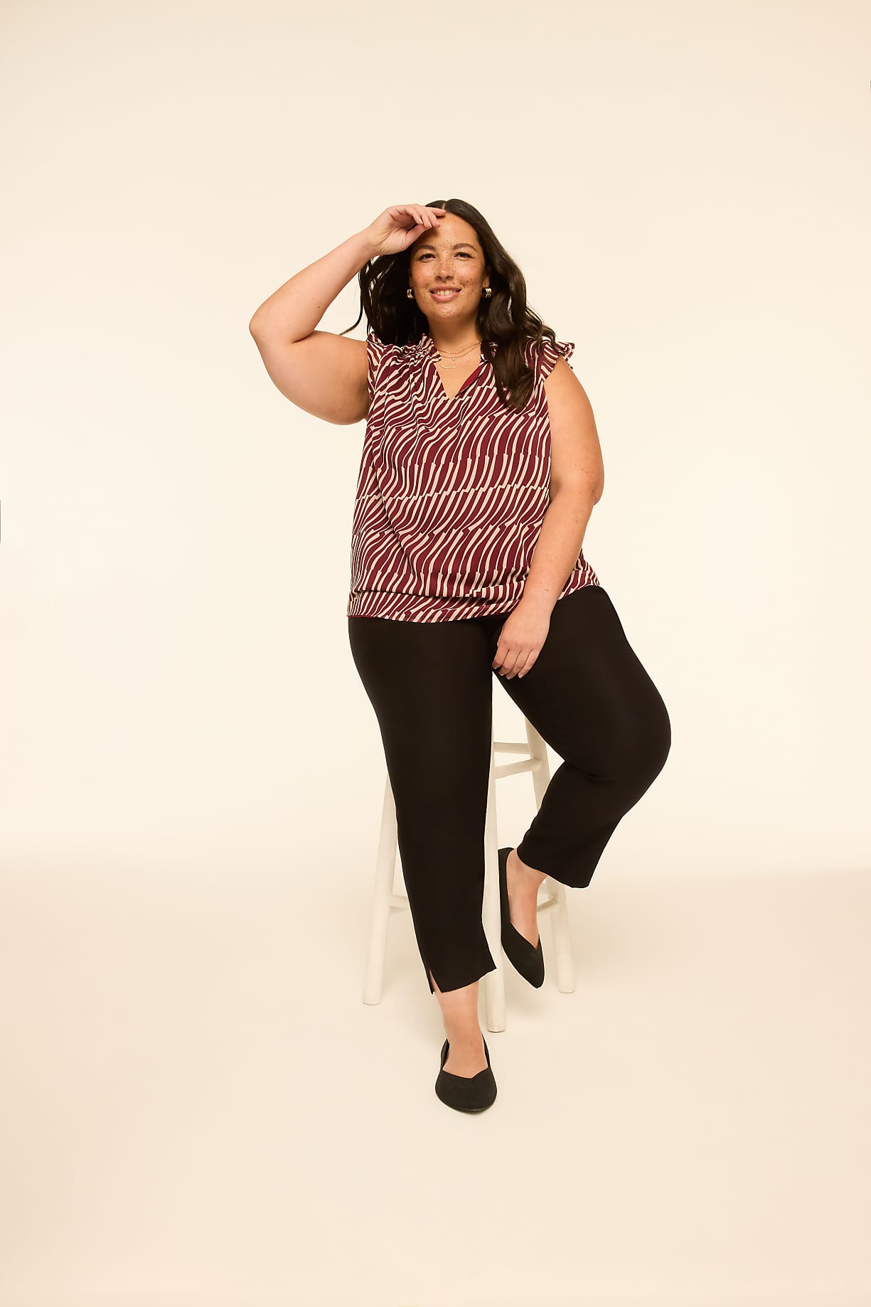 A woman in a patterned top and black pants sits on a stool against a plain, light-colored background. Product Image 5