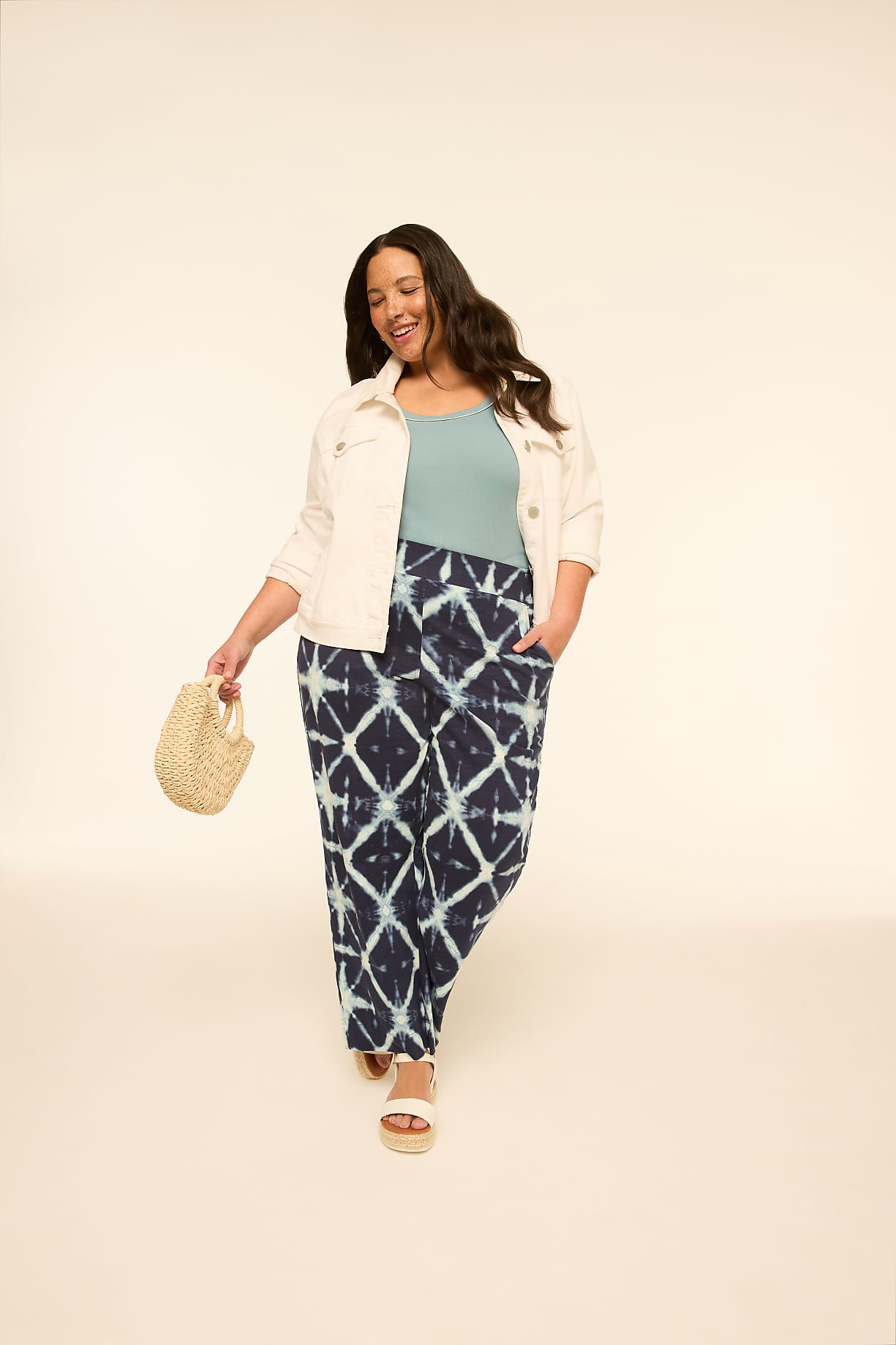 A woman in a light jacket, teal top, and patterned pants walks forward holding a straw bag against a plain, light background. Product Image 6