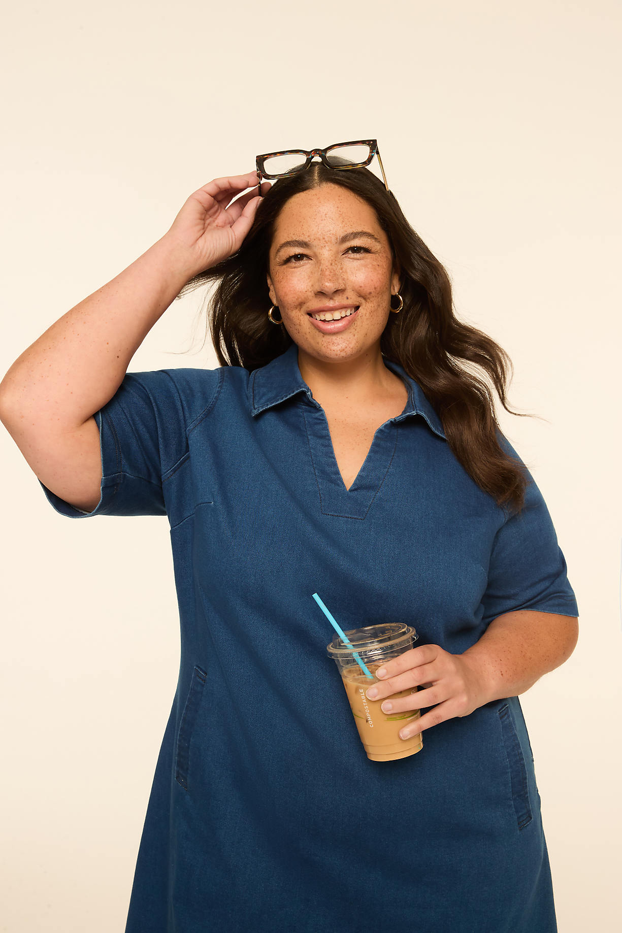 A smiling woman in a denim dress holds a coffee cup while resting glasses on her head against a plain, light-colored background. Product Image 5