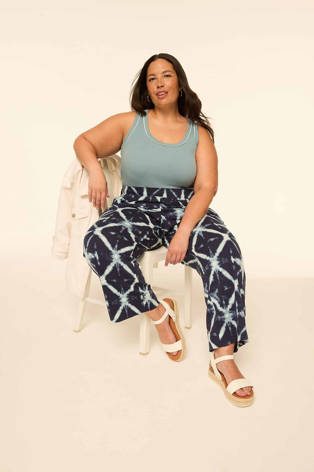A woman with dark wavy hair sits on a white chair in the foreground, wearing a teal tank top and patterned wide-leg pants, with a cream jacket draped over the chair, against a plain, light-colored background. Product Image 7