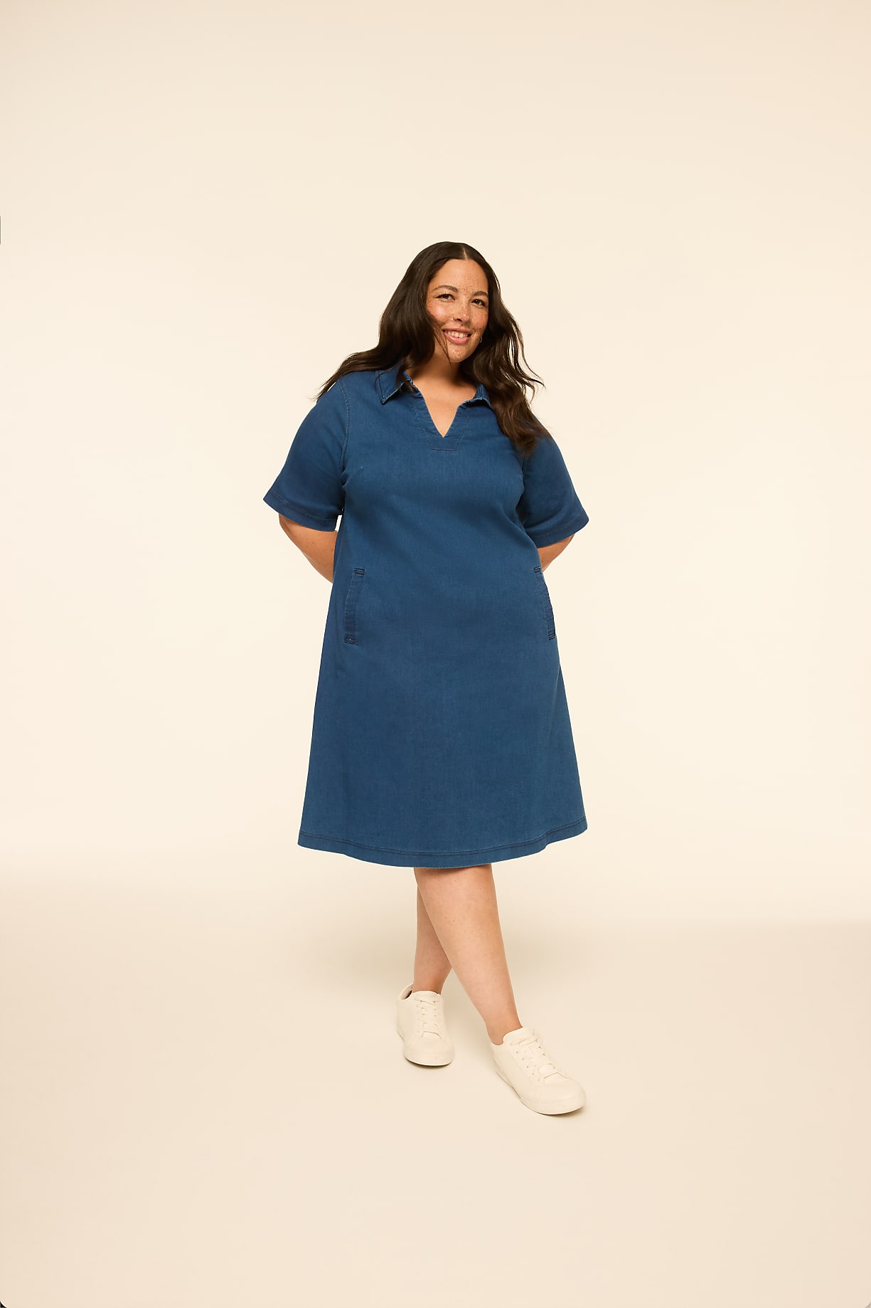 A woman in a blue denim dress and white sneakers stands with her hands behind her back against a plain, light-colored background. Product Image 8
