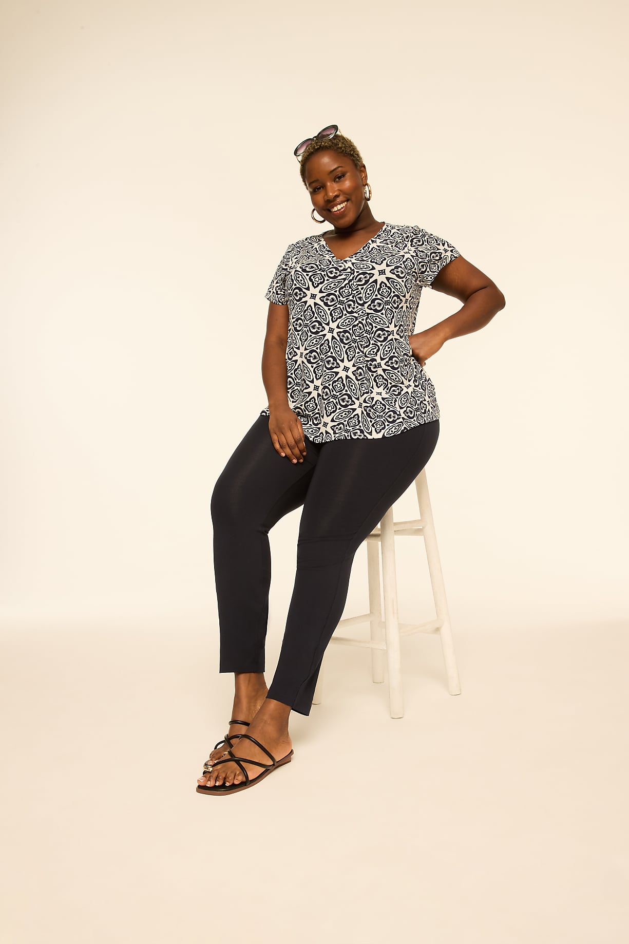A smiling woman in a patterned top and dark pants sits on a stool against a plain, light-colored background. Product Image 7