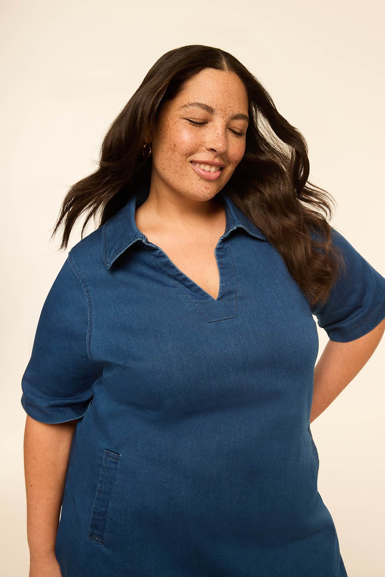 A woman with dark, wavy hair and freckles smiles with her eyes closed, wearing a blue denim dress against a plain, light-colored background. Product Image 9