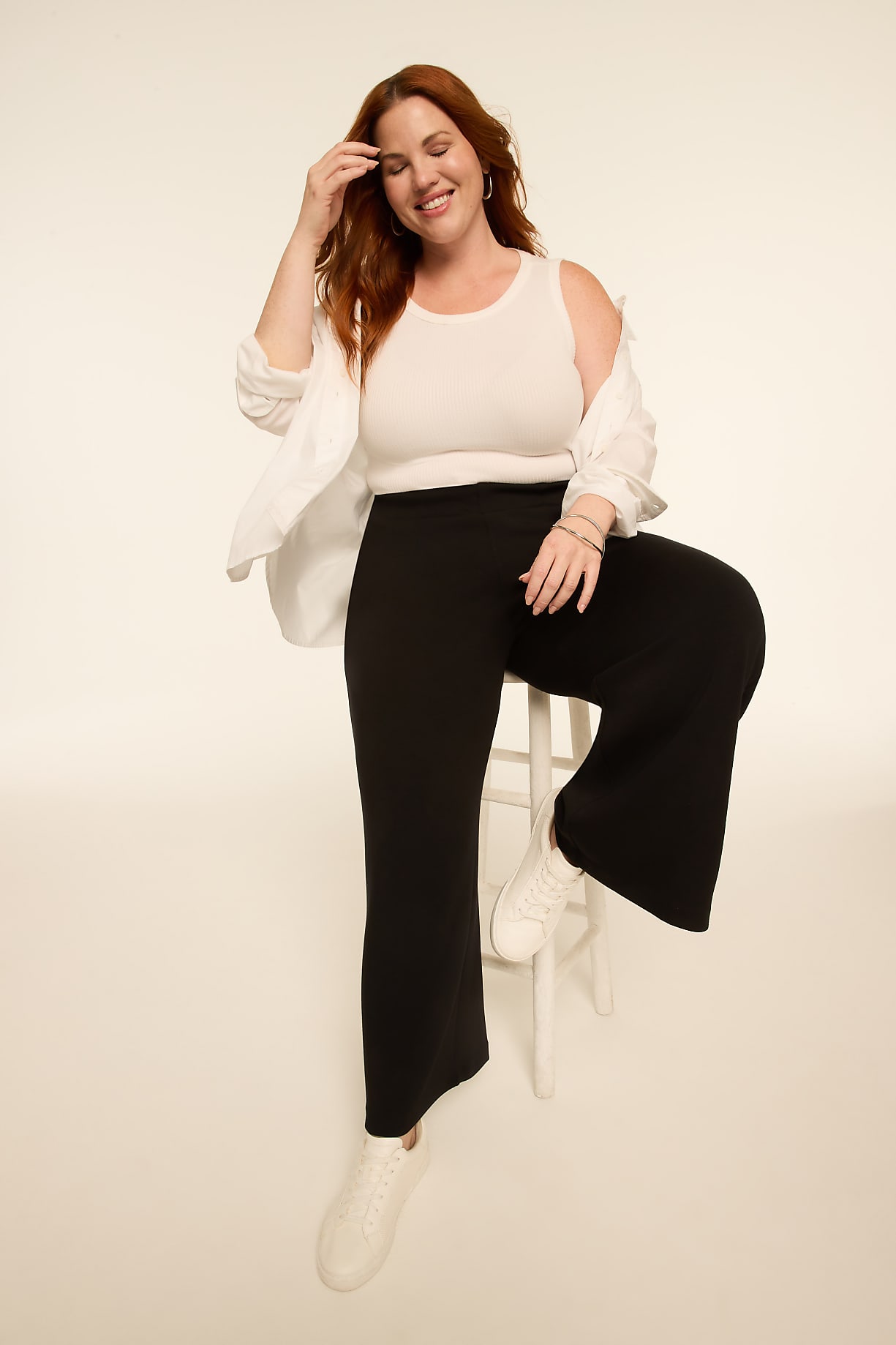 A smiling woman with red hair, wearing a white tank top, black pants, and a white shirt draped over her shoulders, sits on a white stool against a plain, light-colored background. Product Image 6