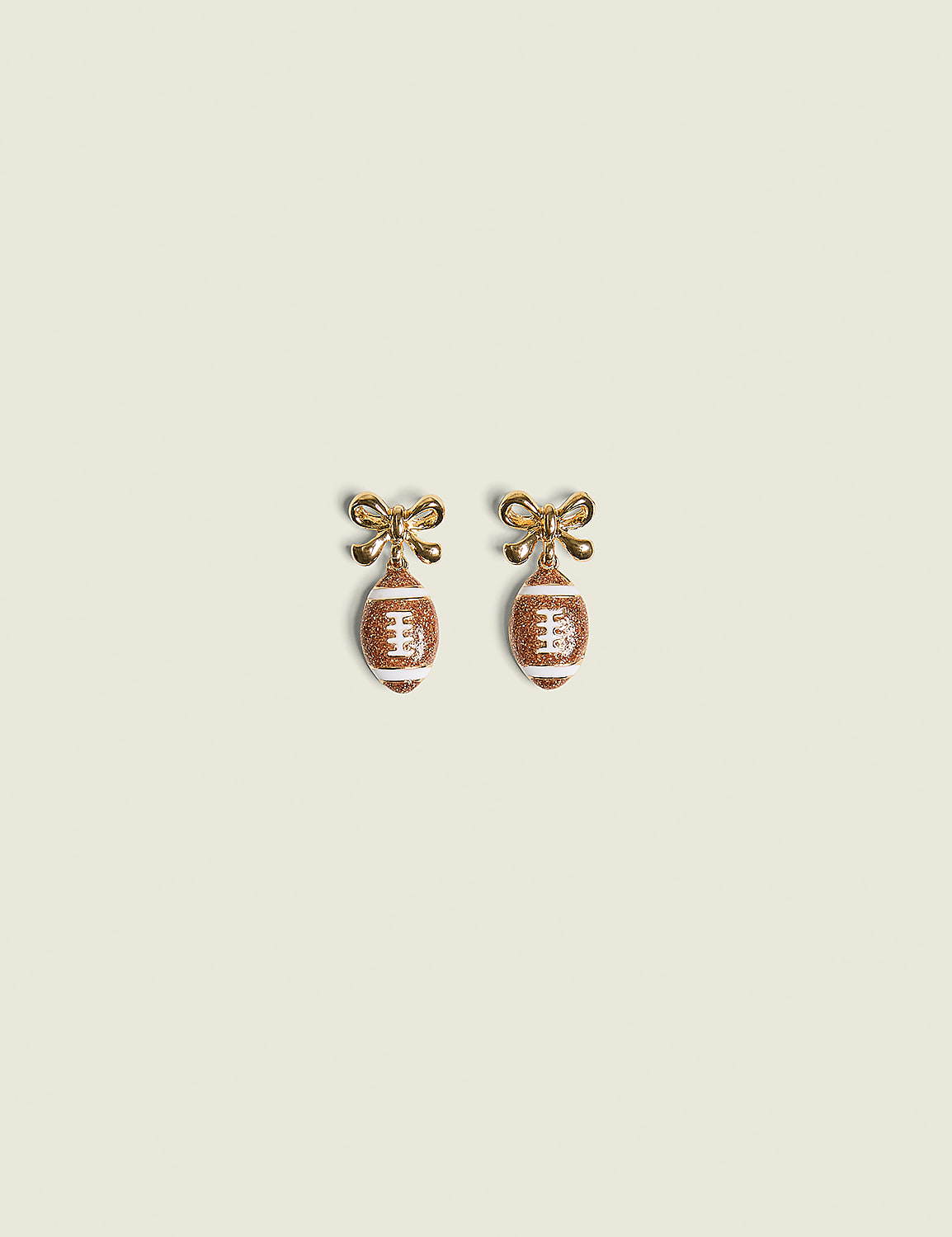 A pair of football-shaped earrings with gold bows hang against a plain, light-colored background. Product Image 1