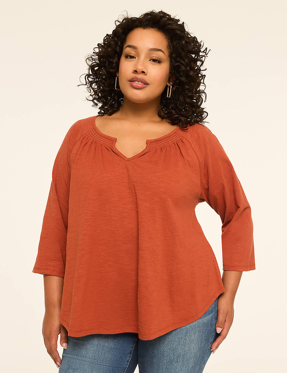 A woman with curly dark hair and a warm smile poses in a rust-colored top and blue jeans against a plain, light-colored background. Product Image 1