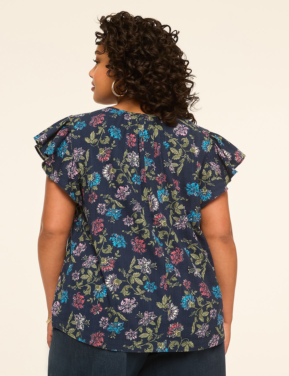 A woman with curly dark hair stands facing away from the camera, wearing a dark blue floral print top with ruffled sleeves, against a plain, light beige background. Product Image 2