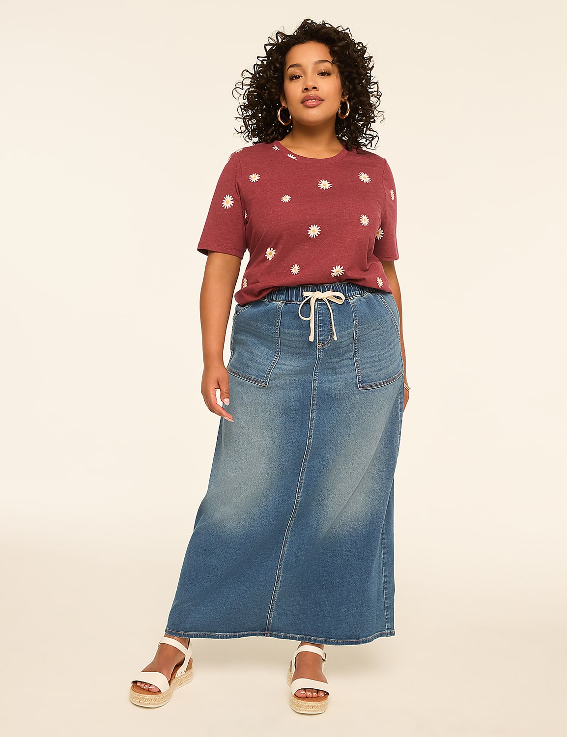 A woman with curly hair stands in a denim maxi skirt and a floral t-shirt against a plain, light-colored background. Product Image 3
