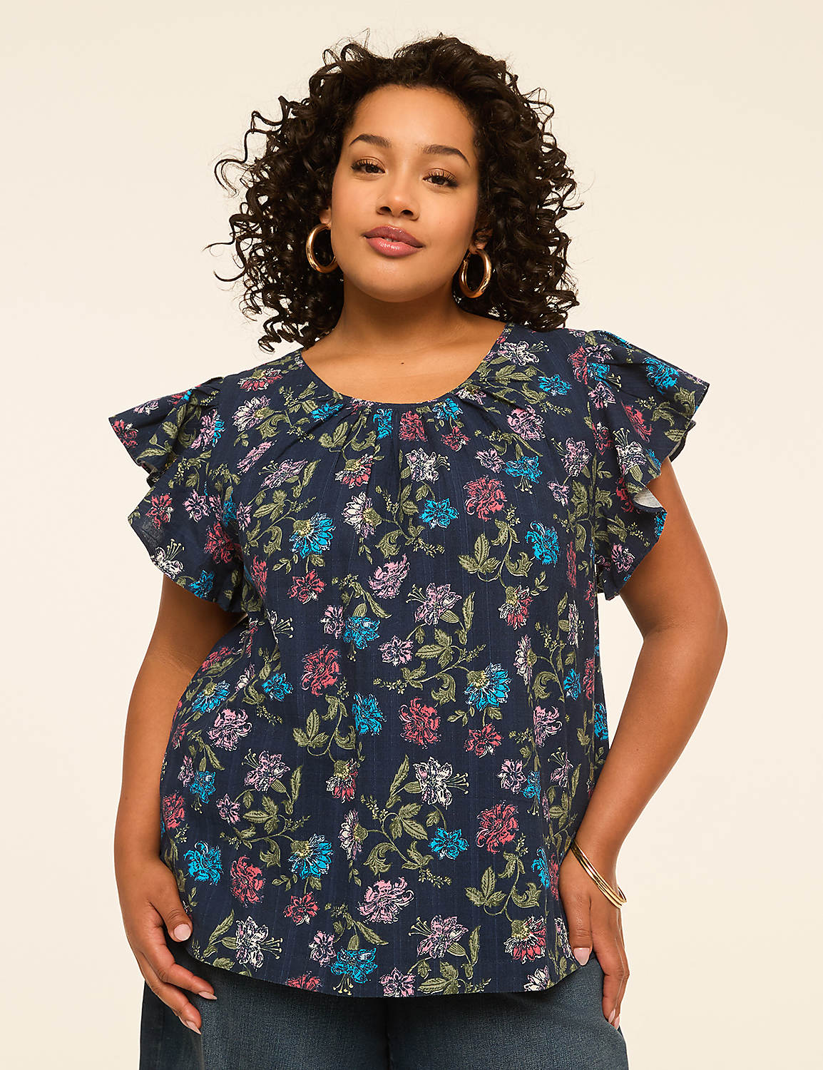 A woman with curly dark hair and hoop earrings poses in a dark blue floral blouse with ruffled sleeves against a plain, light-colored background. Product Image 1