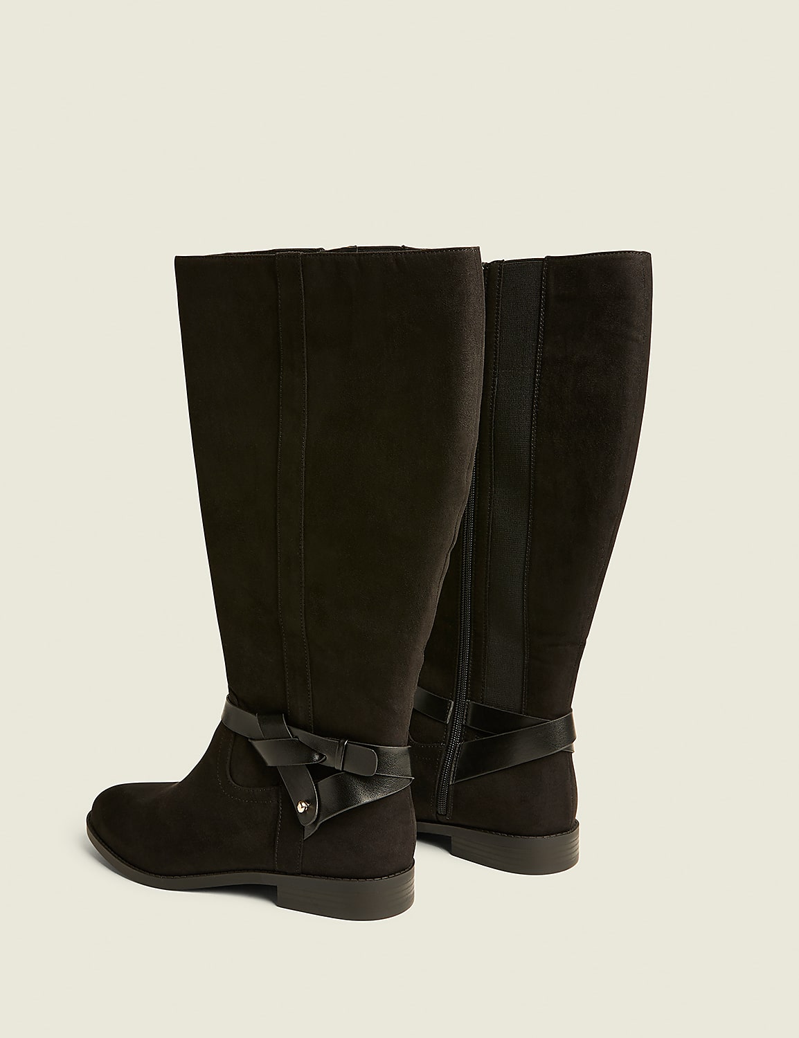 A pair of black suede knee-high boots with a decorative strap and bow in the foreground stand against a plain, light-colored background. Product Image 2