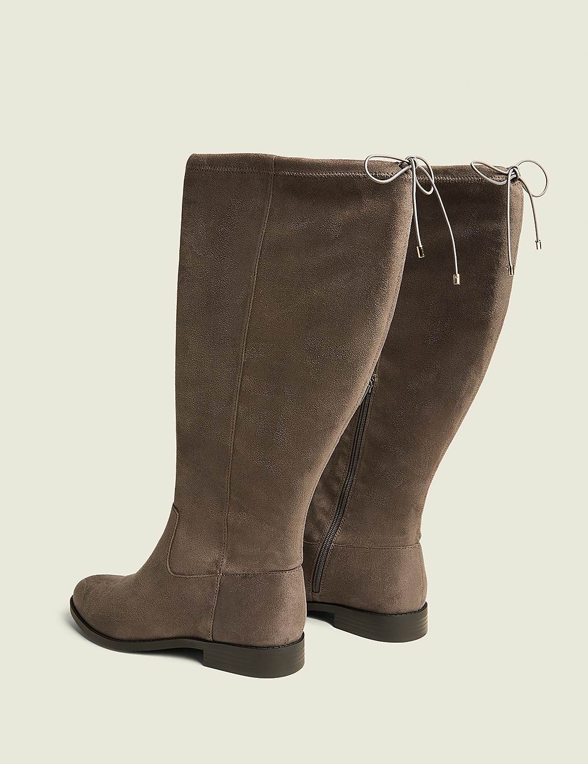 A pair of taupe, knee-high boots with tie details at the back are displayed against a plain, light beige background. Product Image 2