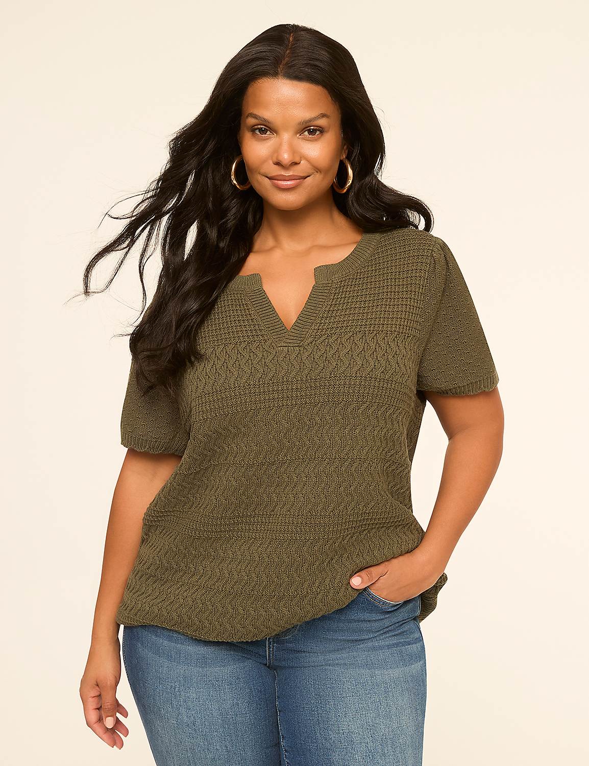 A woman with dark hair and a warm smile stands in the foreground wearing a textured olive green top and blue jeans, against a plain, light beige background. Product Image 1