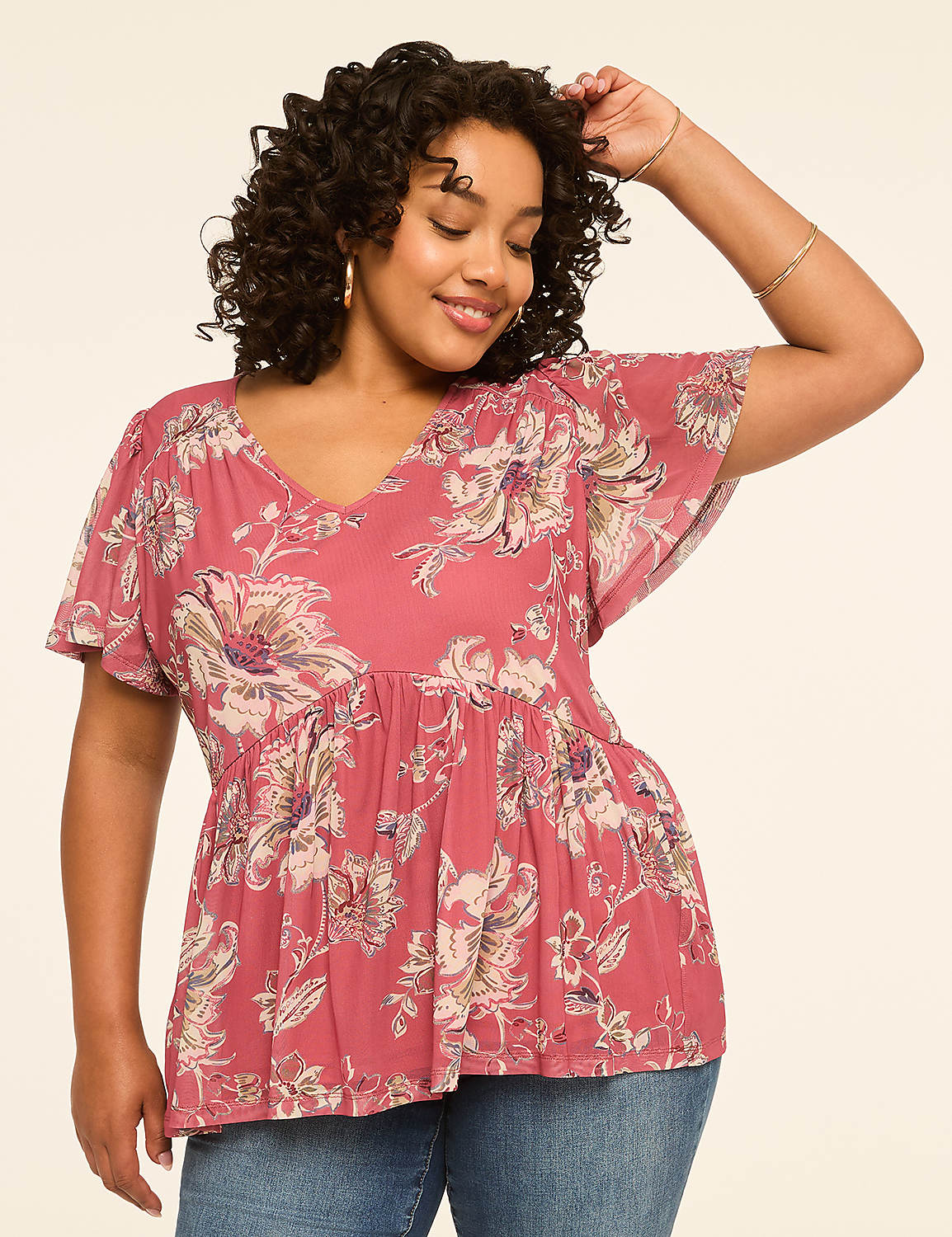 A woman with curly dark hair smiles as she poses in a floral print top and jeans against a plain, light-colored background. Product Image 1