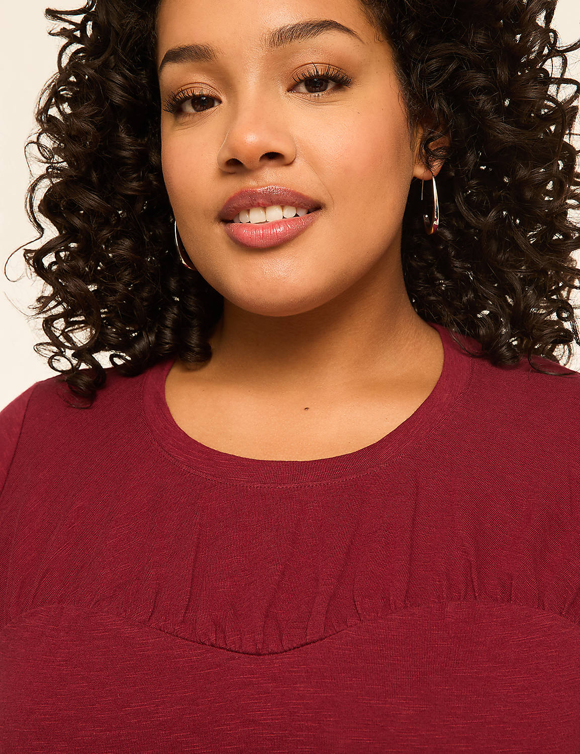 A woman with curly dark hair and hoop earrings smiles gently in the foreground, wearing a maroon top against a plain, light-colored background. Product Image 4