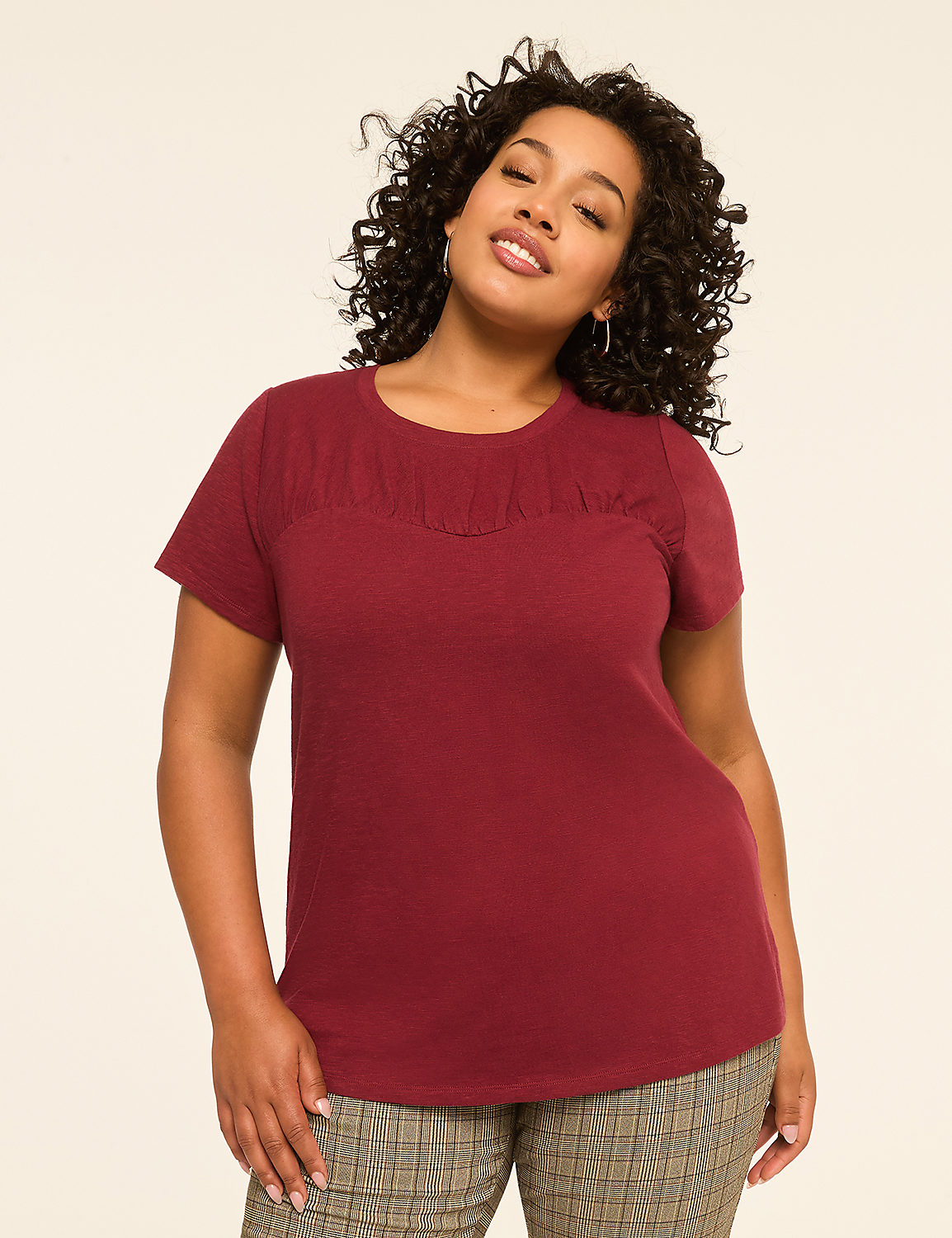 A woman with curly dark hair and a maroon t-shirt stands against a plain, light-colored background. Product Image 1