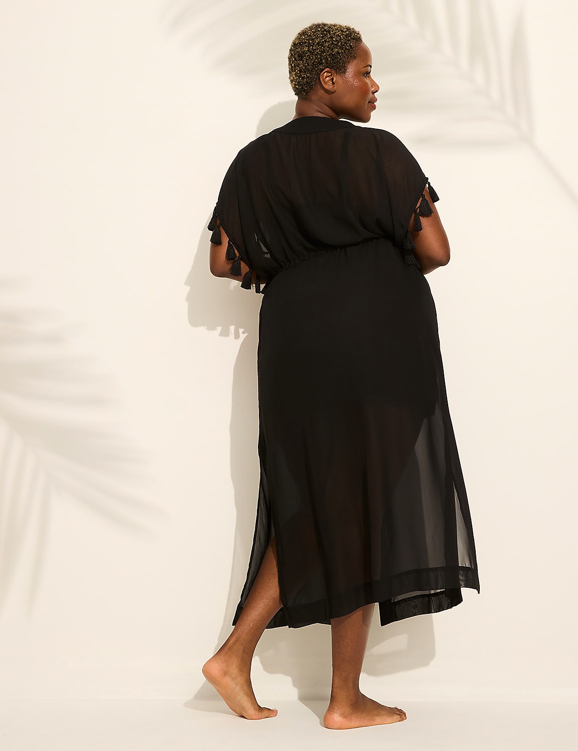 A woman in a black sheer dress stands with her back to the viewer, her bare feet on a white surface, while palm frond shadows are cast on the wall behind her. Product Image 1