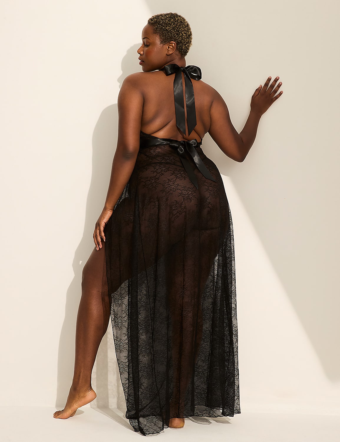 A woman in a sheer black negligee stands with her back to the viewer, leaning against a plain, cream-colored wall. Product Image 1