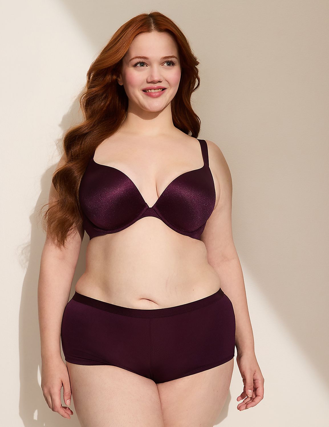 A smiling woman with long red hair poses in a plum-colored bra and matching boy shorts against a softly lit, off-white background with a shadow cast from the left. Product Image 5