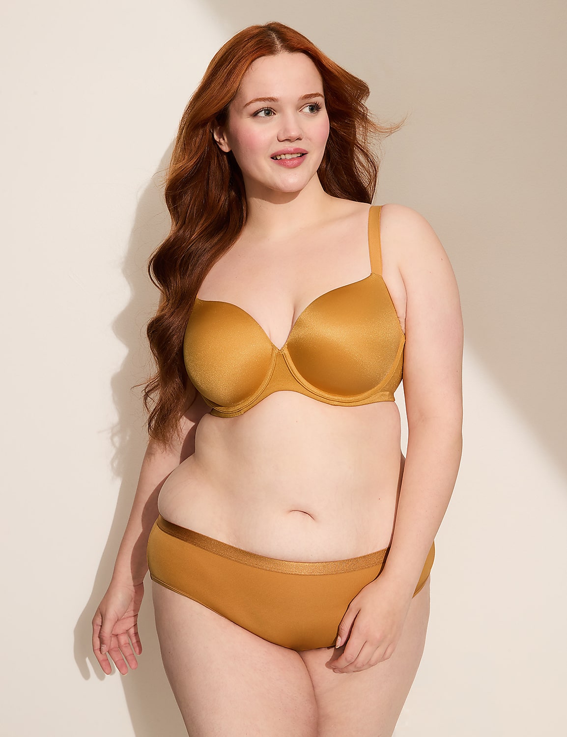 A woman with auburn hair poses in a golden bra and underwear against a plain, softly lit background. Product Image 4