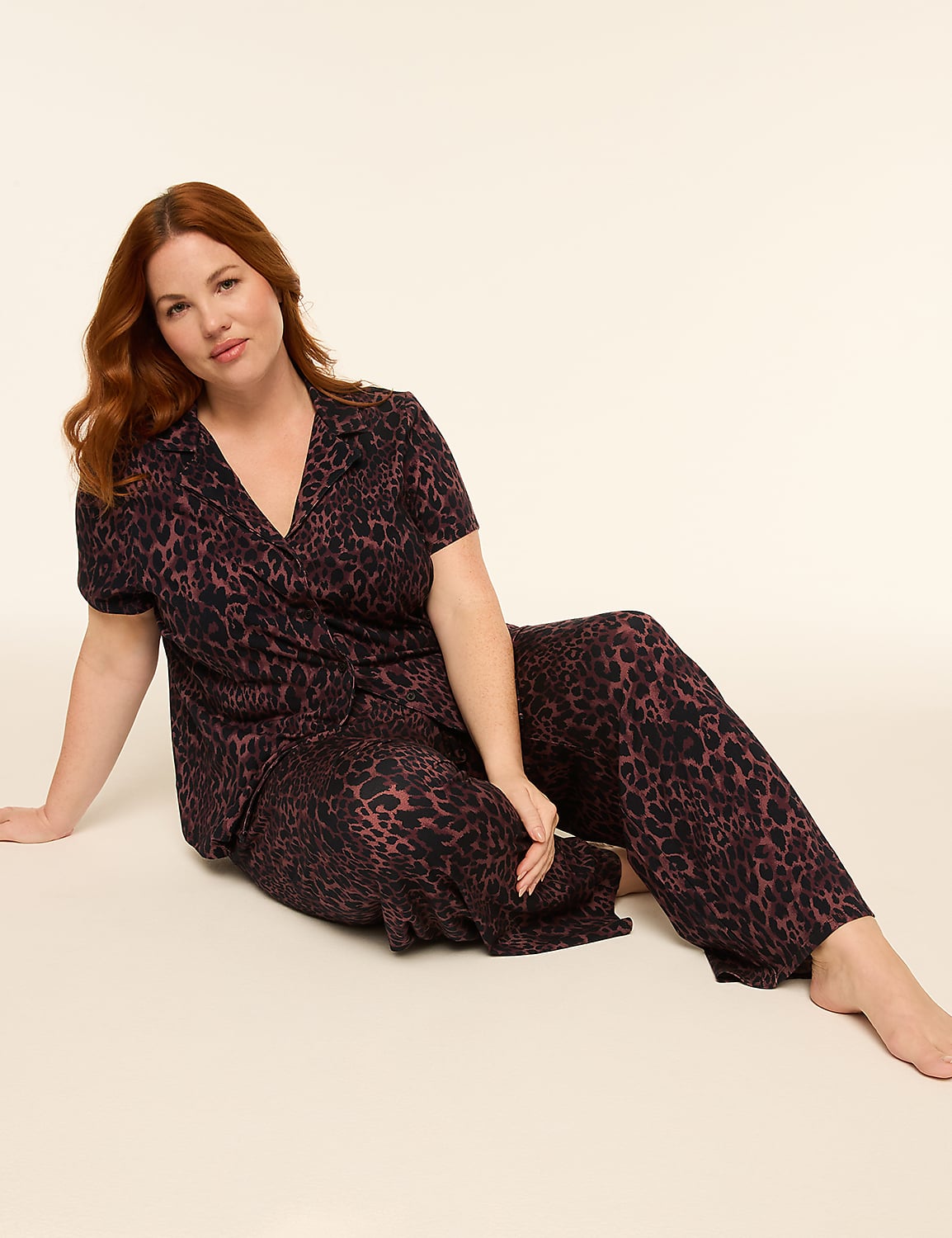 A woman with red hair sits on a light-colored floor wearing a leopard-print pajama set, with a plain, light background behind her. Product Image 1