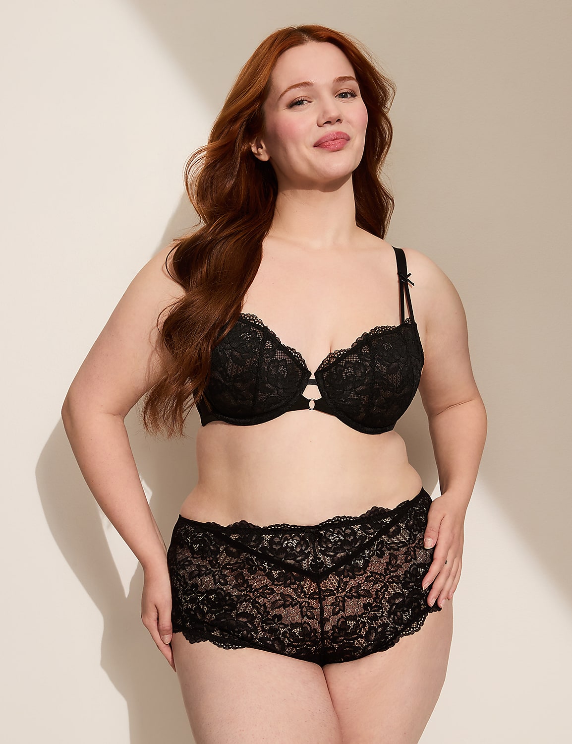 A woman with long red hair poses in black lace lingerie against a plain, light-colored wall with a shadow cast on it. Product Image 4