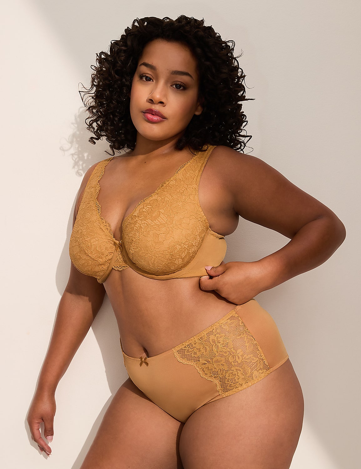 A woman with curly dark hair poses in a tan lace bra and matching underwear against a plain, light-colored wall. Product Image 4