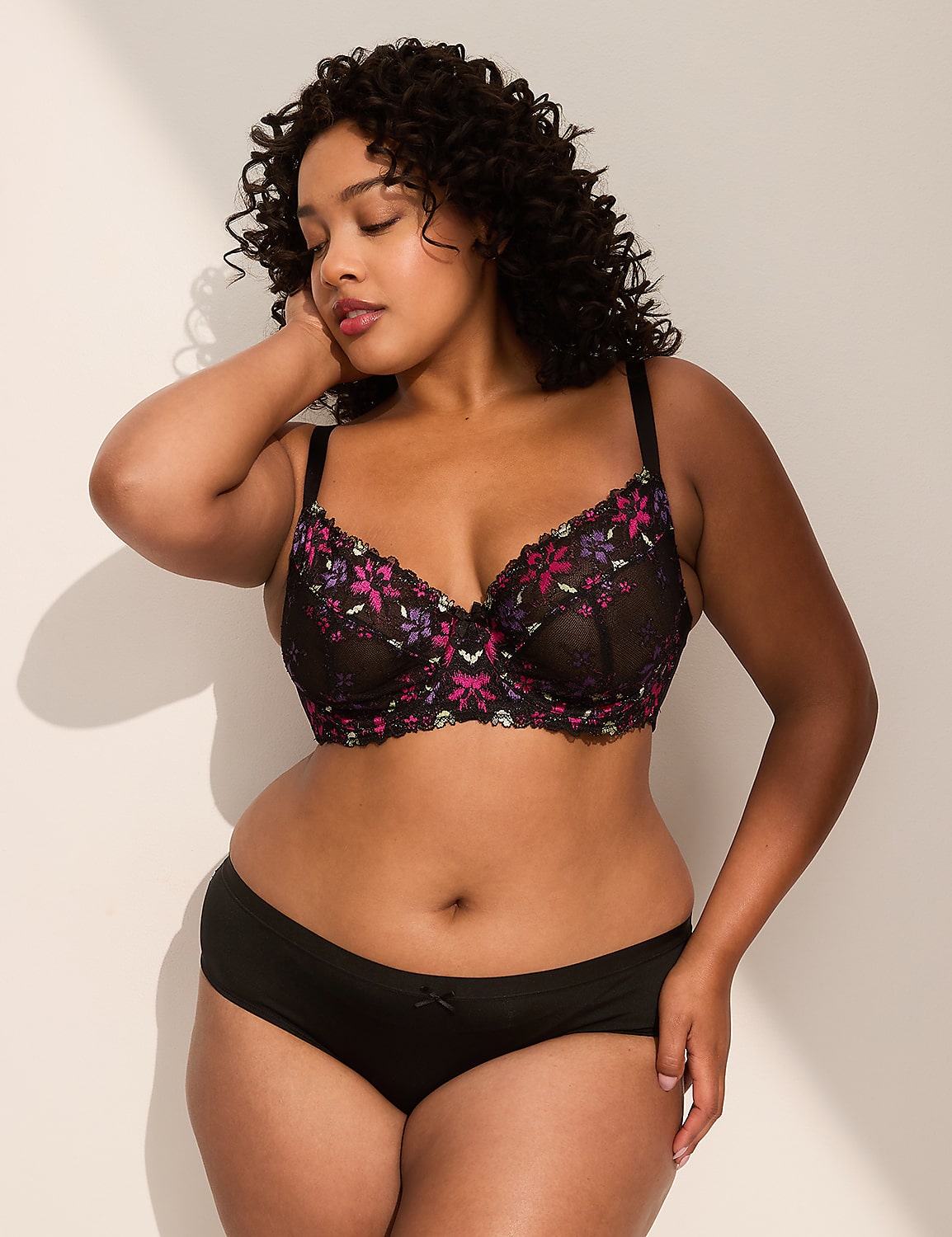 A woman with curly dark hair poses in a black floral bra and black underwear against a plain, light-colored wall with shadows. Product Image 4