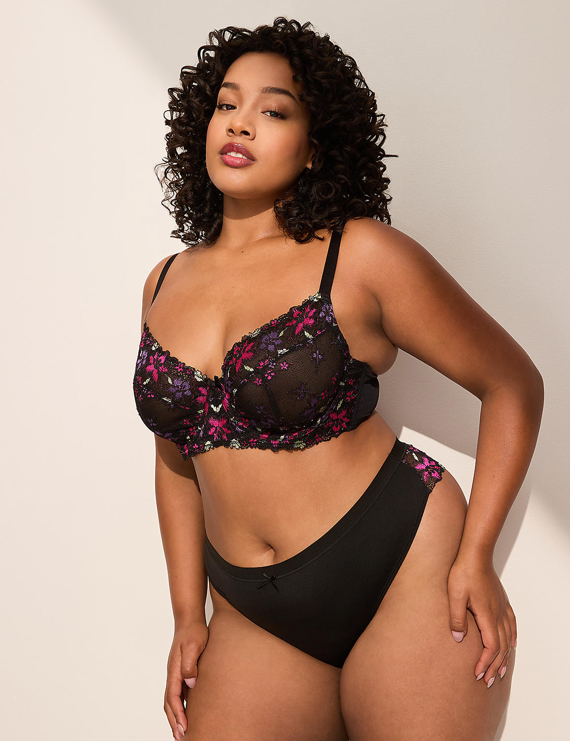 A woman with curly dark hair poses in black floral embroidered lingerie against a plain, light-colored background with soft shadows. Product Image 4