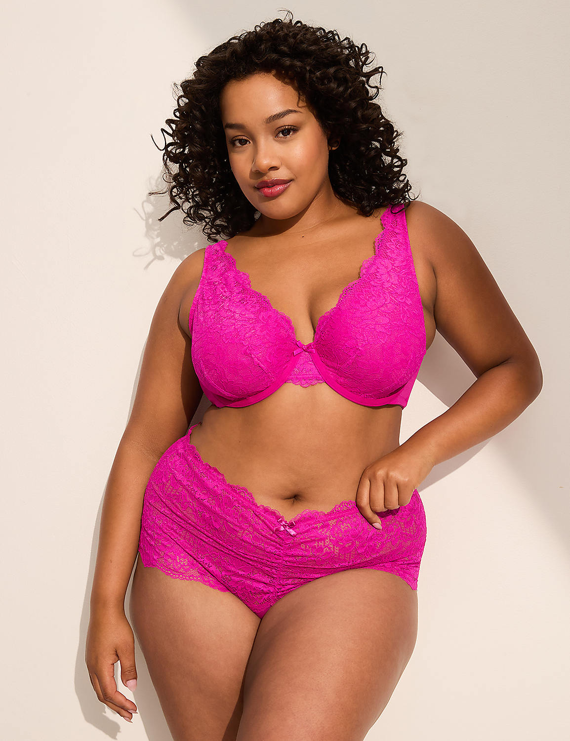 A woman in a pink lace bra and matching underwear stands against a plain, light-colored wall with soft shadows. Product Image 4