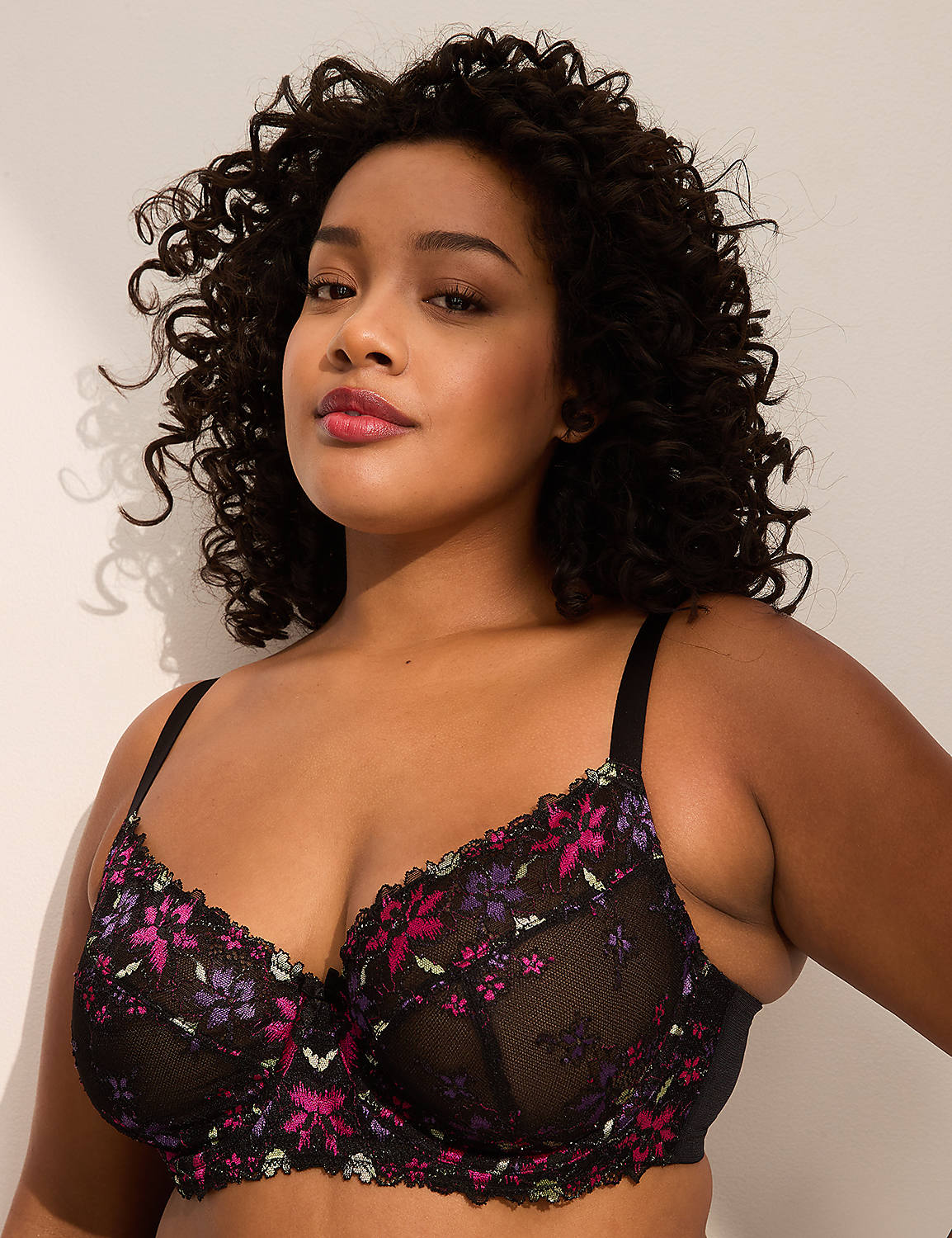 A woman with curly dark hair and a floral lace bra is posed against a plain, light-colored wall. Product Image 1